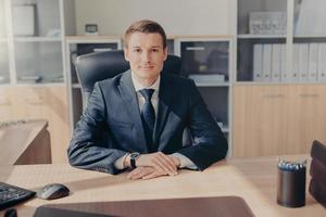 Portrait of attractive prosperous businessman wears black suit, white shirt with tie, sits in his own cabinet, waits for business partners to start meeting, discussing contract terms. Office interior photo