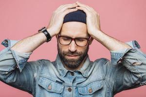 Isolated shot of unhappy desperate man with stubble keeps hands on head, wears fashionable clothing denim jacket, hat and eyewear, poses against pink background. Forgetful male poses indoor photo