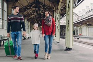 Happy brunette woman wears red sweater and scarf, small kid in oversized jumper, handsome man carries suitcase, return from trip together, express positive emotions. People, family, arrival concept photo