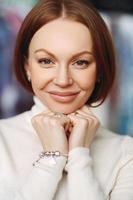 Vertical shot of pleasant looking lady with brown hair, keeps hands under chin, has healthy skin, make up, wears bracelet and white jumper, looks at camera, has broad forehead, nice appearance photo