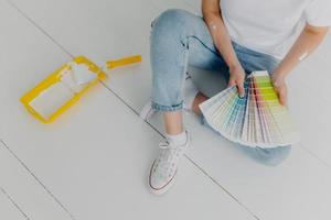 Faceless woman in jeans holds color samples, chooes best tone for refurbishment walls, sits on white floor, uses tray and paint roller for house remodeling. Home makeover and renovation concept photo