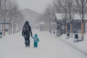 Outdoor shot of small child and father cover distance, being on way to home, hold hands, enjoy winter snowy weather. Back view of people walking on street during snowfall. Cold season concept photo