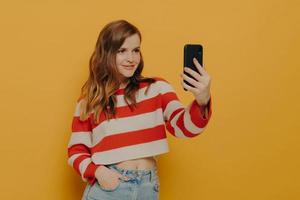 Young charming woman blogger talking selfie on mobile phone while posing on yellow background photo