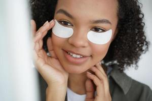 Young biracial girl applying patches on under eye skin smiling. Morning beauty routine, skincare photo