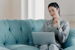 Serious woman entrepreneur takes off glasses, focused in laptop computer screen, analyzes statistics data, looks through financial information, works online, sits on sofa in coworking space. photo