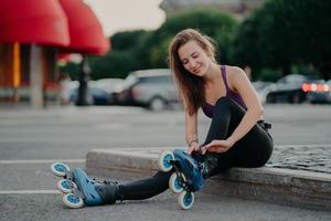 People leisure fitness sport recreation concept. Pleased young woman puts on rollerskates going to ride rolles in urban place has regular exercising goes in for dangerous sport adjustes laces photo