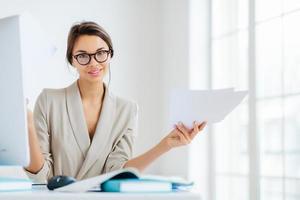 Indoor shot of good looking female in formal suit, holds paper documents, works on organization and creating startup project, poses at desktop with computer, checks information for presentation photo