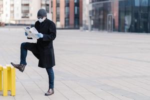 Sick man with flu wears medical mask to protect spreading infectious disease, dressed in coat, stands at street near office building, reads newspaper, finds out news about coronavirus pandemic photo