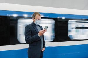 Horizontal shot of male traveler uses public transport for commuting, wears medical mask to protect from coronavirus or covid-19, waits for train, uses mobile phone, sends text messages in chat photo