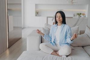 Woman practice yoga, meditating, breathing, relaxing in lotus pose on couch at home. Stress relief photo