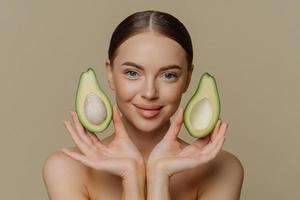 Personal care and spa concept. Brunette satisfied European woman holds halves of avocado near face recommends face mask stands with bare shoulders indoor isolated over beige studio background. photo