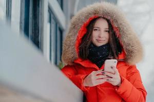 Outdoor shot of lovely dark haired Caucasian woman in winter street clothes, holds modern cell phone, reads received message, surfes social networks, enjoys spare time and snow, chilly weather photo