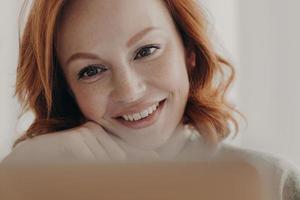 Close up shot of positive redhead woman with freckled skin and toothy smile, concentrated at display of laptop computer, satisfied with online business, checks received message, has remote job photo