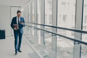 Smiling redhead man office worker in suit using cellphone while standing in modern office photo