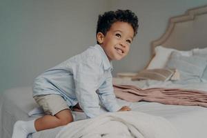 niño afroamericano sonriente tratando de subirse a una cama grande en casa, divirtiéndose en el interior foto