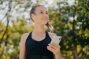 Thoughtful cheerful woman with pony tail has walk outdoor listens music while going in for sport holds modern smartphone leads healthy lifestyle focused into distance poses at park against trees photo