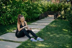 Sporty female blogger talks with followers online talks about healthy lifestyle and fitness training shares media in networks uses wireless earphones dressed in activewear poses against green grass photo