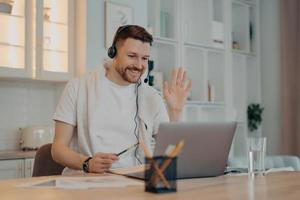 concepto de aprendizaje electrónico. feliz hombre barbudo estudiante tiene videoconferencia estudios en línea ondas palma en la pantalla de la computadora portátil saluda al maestro trabaja a distancia usa auriculares toma notas en el diario posa sobre el interior de la casa foto