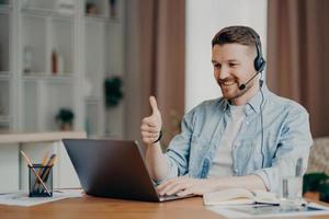 I like it. Glad bearded adult man has video conference keeps thumb up likes something focused at laptop computer has distance job interview dressed casually. Professional call center agent working photo