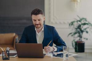 Indoor shot of handsome male office worker rewrites information from notepad in diary sits at desk dressed in formal clothes spends time for searching info. Businessman takes notes does paper work photo