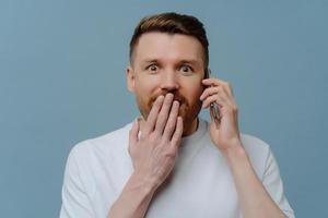 Astonished surprised bearded adult man covers mouth looks with amazement at camera has telephone conversation keeps smartphone near ear isolated over blue background. Human reactions concept photo