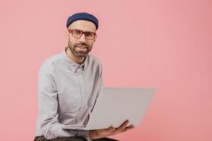 Unshaven male blogger updates his profile, surfes social networks, shares multimedia files with followers, connected to wireless internet, holds laptop computer, wears stylish formal clothes photo
