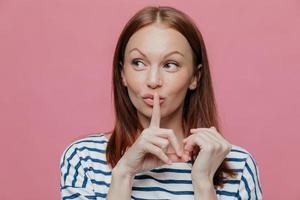 Pretty curious Caucasian woman makes silence sign, keeps index finger over mouth, looks secretly aside, wears striped casual clothes, poses over pink studio wall with free space for your text. Hush photo