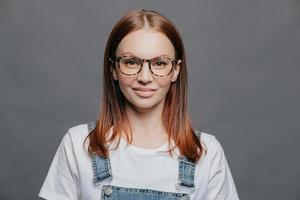 Happy young woman with glad facial expression, has healthy skin, wears optical glasses, white t shirt and denim sarafan, models over grey background, going to meet with friends, poses indoor photo