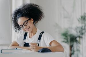 Happy Afro American female student writes down information, prepares for exams, sits in coworking space, has curly dark hair, wears optical glasses white t shirt and overalls, studies in spacious room photo