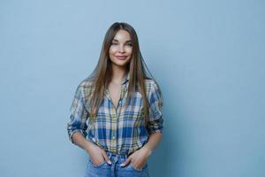 Attractive student glad to talk about part time job, keeps hands in pockets of jeans, being happy because of positive news, has pleasant smile, isolated over blue studio background. People and style photo