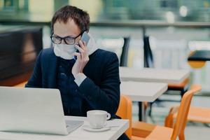 Serious busy man worker works in quarantine, reads information online on laptop computer, wears protective medical mask, calls someone via cellular, cares about health during epidemic virus infection photo