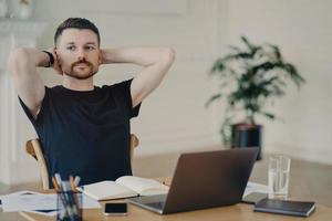 Pensive male freelancer takes break keeps hands behind head poses at desktop works on laptop computer has thoughtful expression thinks about future plans plans week dressed in black t shirt. photo