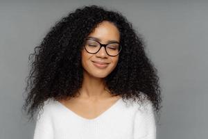 Close up shot of pleased smiling woman with bushy crisp hair, keeps eyes shut, smiles gently, wears optical glasses, imagines something pleasant, dressed in white jumper, isolated over dark grey wall photo