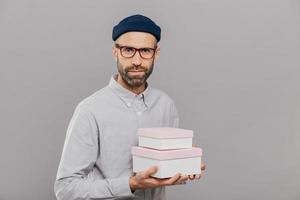 Photo of serious male model dressed in festive clothes, holds gifts in hands, going to congratulate friend with birthday, ready for party wears spectacles isolated over grey wall. This present for you