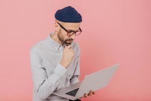 Experienced male freelancer develops new strategy, holds modern laptop computer, keeps hand on chin, wears formal clothing, busy with distant work, isolated over pink background, copy space. photo