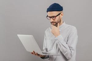 Attentive student holds chin, focused in monitor of laptop computer, searches information for project, connected to wireless internet, isolated over grey studio wall. Networking and people concept photo