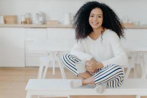 Smiling relaxed African American female sits crossed legs on bench against kitchen interior, wears white sweater and striped pants, drinks hot beverage, enjoys domestic atmosphere. Coffee time photo