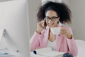 Photo of attractive smiling female entrepreneur enjoys aromatic beverage, holds white mug of drink, has telephone conversation, dressed in formal outfit, poses at workplace, wears transparent glasses