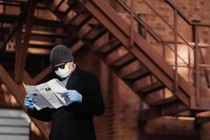 Coronavirus precautions. Male model avoids crowded places, fights against new virus, reads attentively article about covid-19 symptoms, holds newspaper, wears protective face mask and medical gloves photo