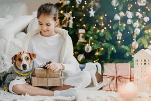 Photo of lovely small child sits crossed legs on floor, pets pedigree dog received Christmas presents from parents celebrate New Year together. Kid enjoys holiday with jack russell terrier opens gifts