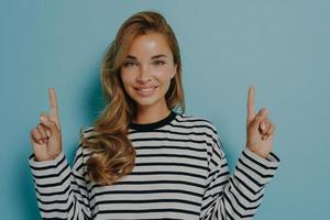 Photo of lovely smiling young woman points fingers up shows promo logo overhead