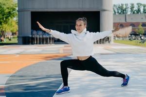 la deportista de ajuste flexible hace ejercicios deportivos al aire libre  mantiene los brazos levantados tiene una expresión alegre usa polainas con  capucha y entrenadores posa en el estadio en un lugar