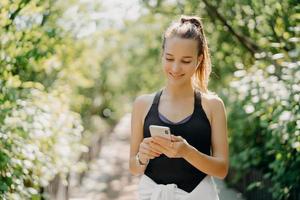 Sport lifestyle and workout concept. Young pleased sporty woman uses smartphone for chatting online and listening music rests after cardio training has combed pony tail dressed in active wear photo