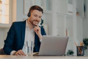 Professional male operator works in callcenter uses headset and laptop computer talks on video call or virtual webcam event wears formal clothes poses against cozy interior has online meeting photo