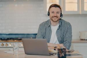 Handsome glad man customer support manager gives advice or consultation to clients works online from home poses at home kitchen wears wireless headphones uses modern laptop computer. Remote job photo