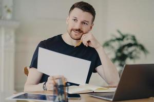 Glad handsome European businessman holds paper document poses in home interior at desktop surrounded by modern technologies checks information has remote work looks thoughtfully into distance photo