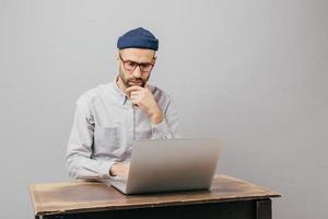 Successful male designer keyboards on laptop computer, checks database, sits at old table, holds chin, wears formal shirt, spectacles for vision correction, isolated over grey background indoor photo