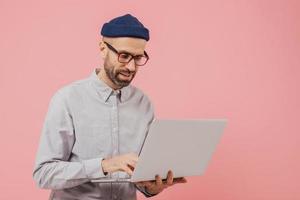 Professional male worker searches for interesting movie to watch, uses gadget, types information on laptop computer, wears formal clothes, isolated over pink background. Online communication photo