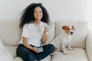 Positive lovely teenage girl with glad expression, texts message in social media, uses app on cellular, connected to wireless internet, poses on couch with dog, drinks tea, spends free time at home. photo