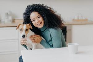 Adorable smiling woman embraces her favourite dog, expresses care and love, good attitiude to pet, poses against kitchen interior, drinks aromatic beverage. People, animals, relationship, affection photo